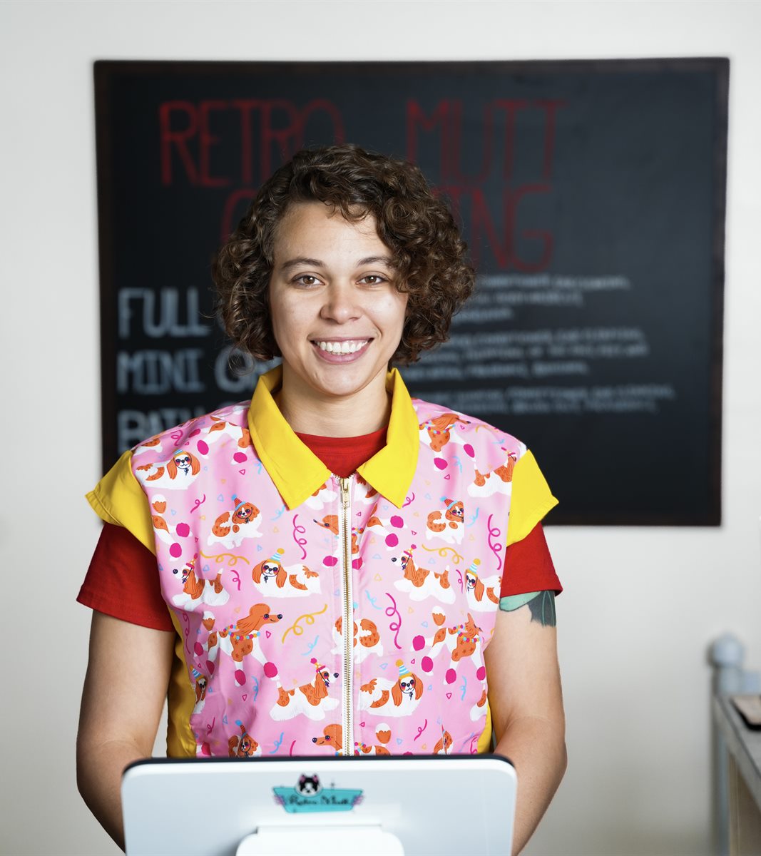A Retro Mutt groomer at the front desk in a patterned grooming smock, service chalkboard on the wall behind